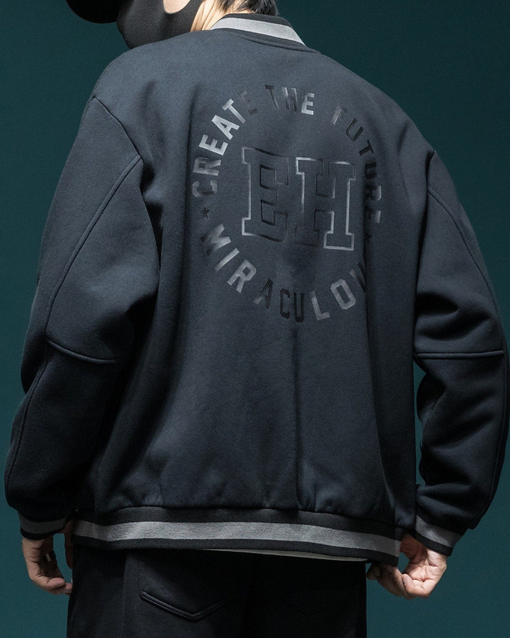Back of the dark varsity jacket with the circular CREATE THE FUTURE, EH, MIRACULOUS print and the striped ribbed hem