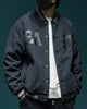 Dark slate-black varsity jacket with EA and ST chest letters, snap front and striped ribbed collar, worn over a white tee