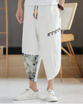 White linen drop-crotch pants with a toggle patch row on the thigh and an ink-wash print panel on the lower leg