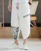 White linen drop-crotch pants with a toggle patch row on the thigh and an ink-wash print panel on the lower leg