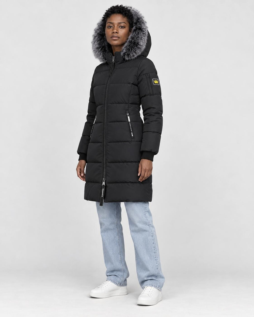 Model wearing the black puffer coat with a dark gray faux-fur hood, light-wash jeans and white sneakers, three-quarter view
