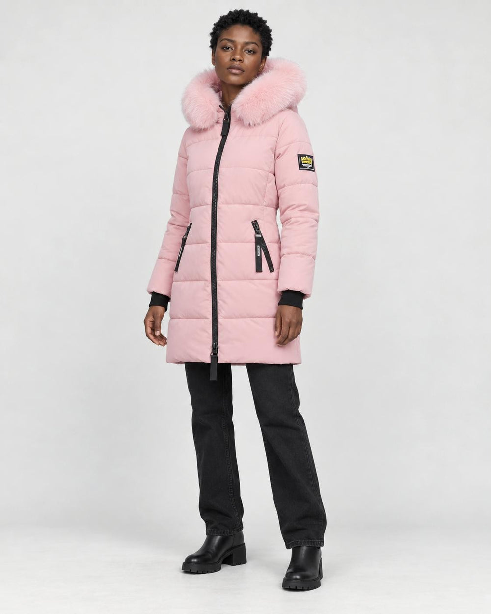 Model wearing the pink long puffer coat with a pink faux-fur hood, black jeans and black boots, three-quarter view