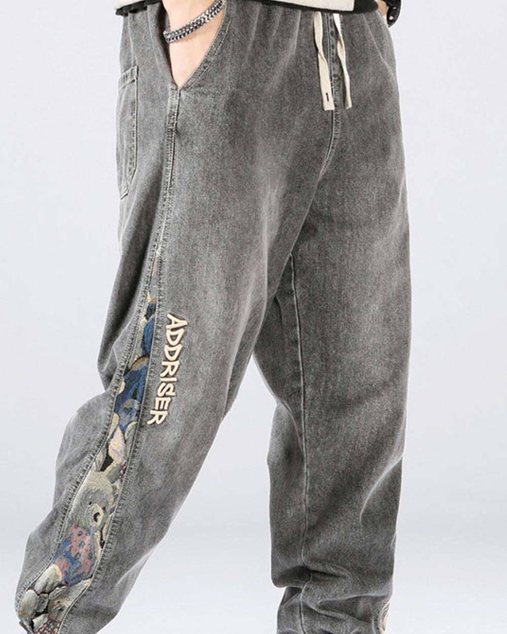 Man in the washed gray denim joggers, showing the woven teddy-bear panel and the ADDRISER lettering down the outer leg