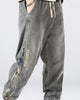 Man in the washed gray denim joggers, showing the woven teddy-bear panel and the ADDRISER lettering down the outer leg