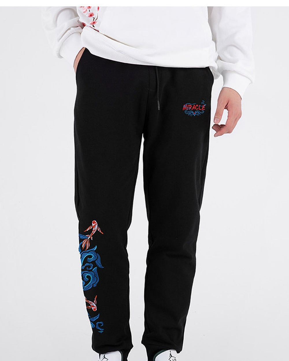 Man in black embroidered jogger pants with a white sweatshirt, front view with a hand in the pocket