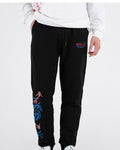 Man in black embroidered jogger pants with a white sweatshirt, front view with a hand in the pocket