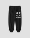 Black cotton jogger pants with a white crossed-out-eyes smiley print on the thigh, laid flat