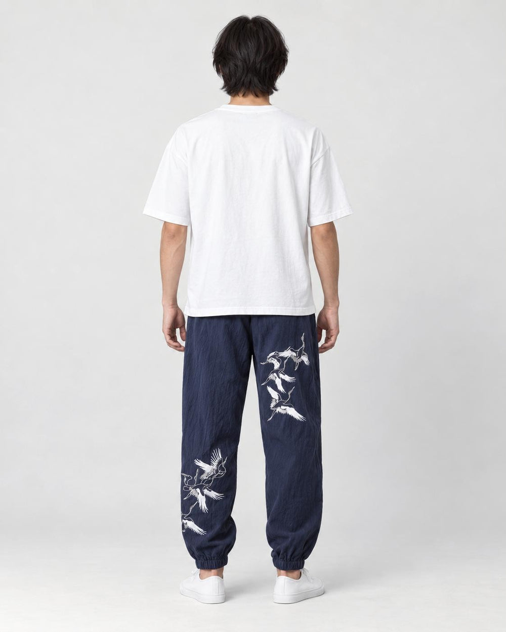 Model seen from behind in the navy crane-embroidered jogger pants, a white tee and white sneakers