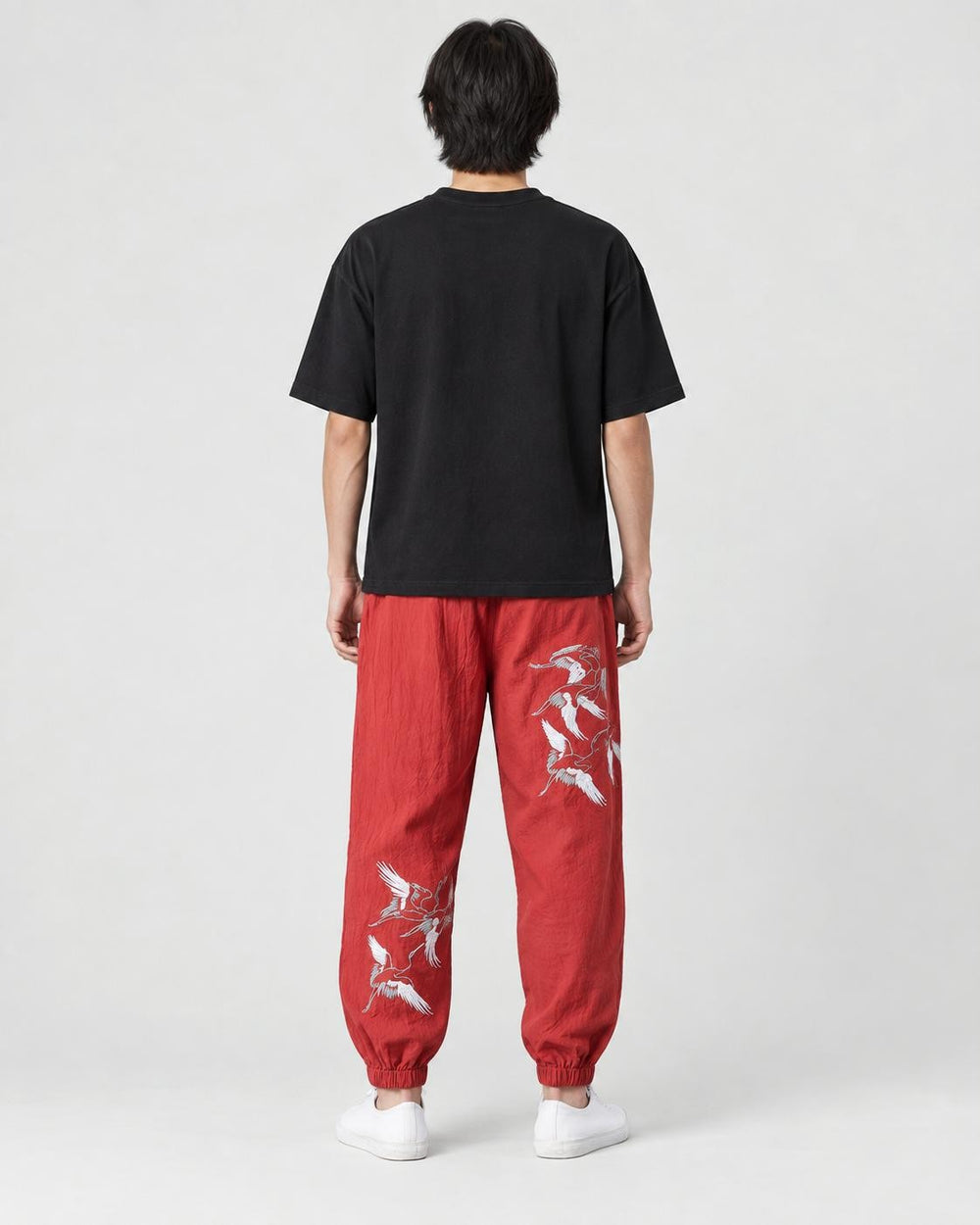 Model seen from behind in the red crane-embroidered jogger pants, a black tee and white sneakers