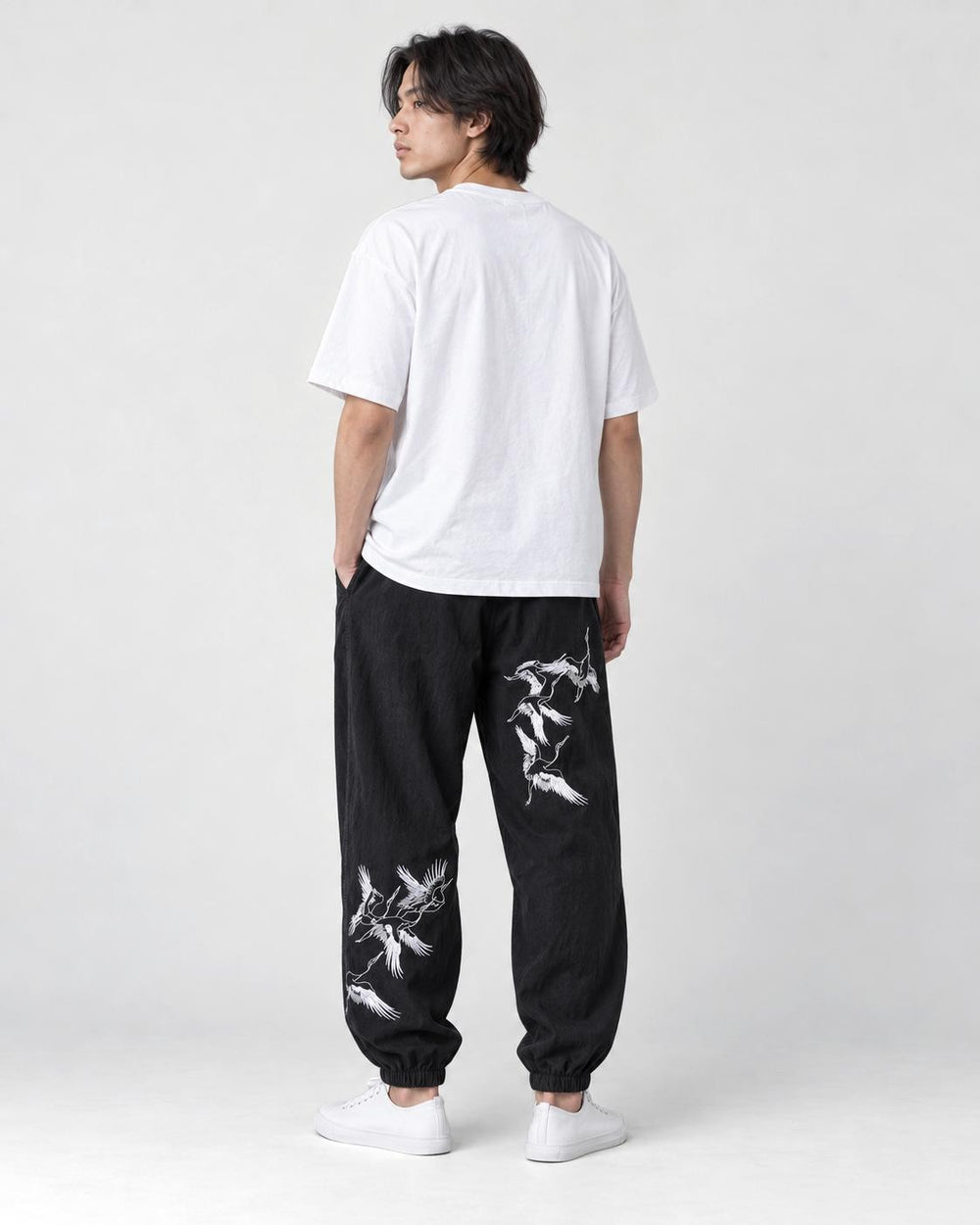 Model seen from behind at an angle in the black crane-embroidered jogger pants, a white tee and white sneakers