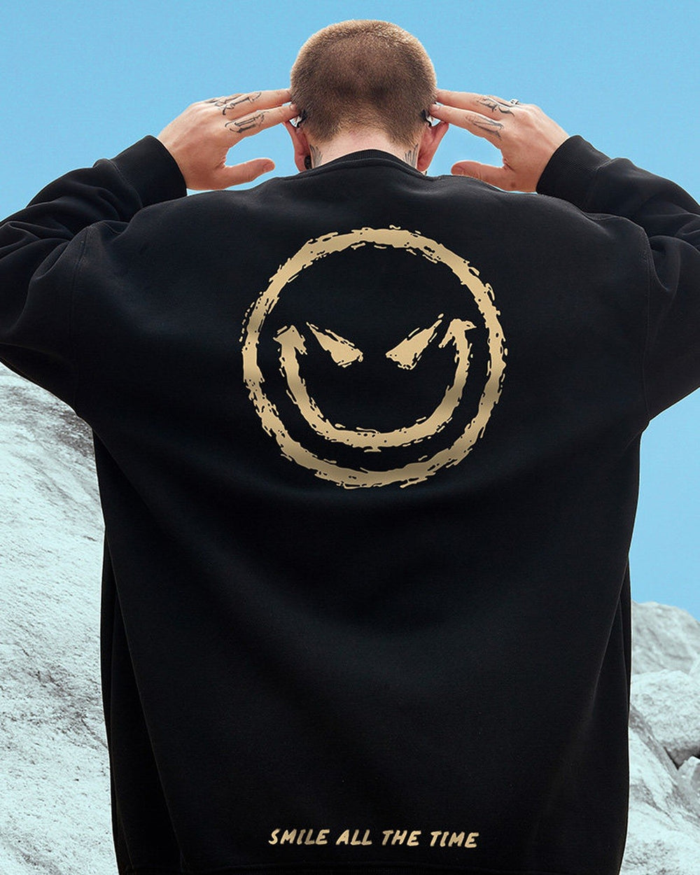 Man with his hands behind his head in the black crewneck sweatshirt, showing the gold devil smiley on the back