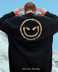 Man with his hands behind his head in the black crewneck sweatshirt, showing the gold devil smiley on the back
