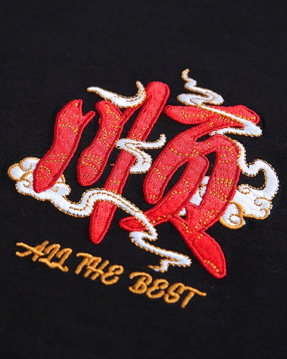 Close-up of the small red character and gold ALL THE BEST lettering embroidered on the chest of the black T-shirt