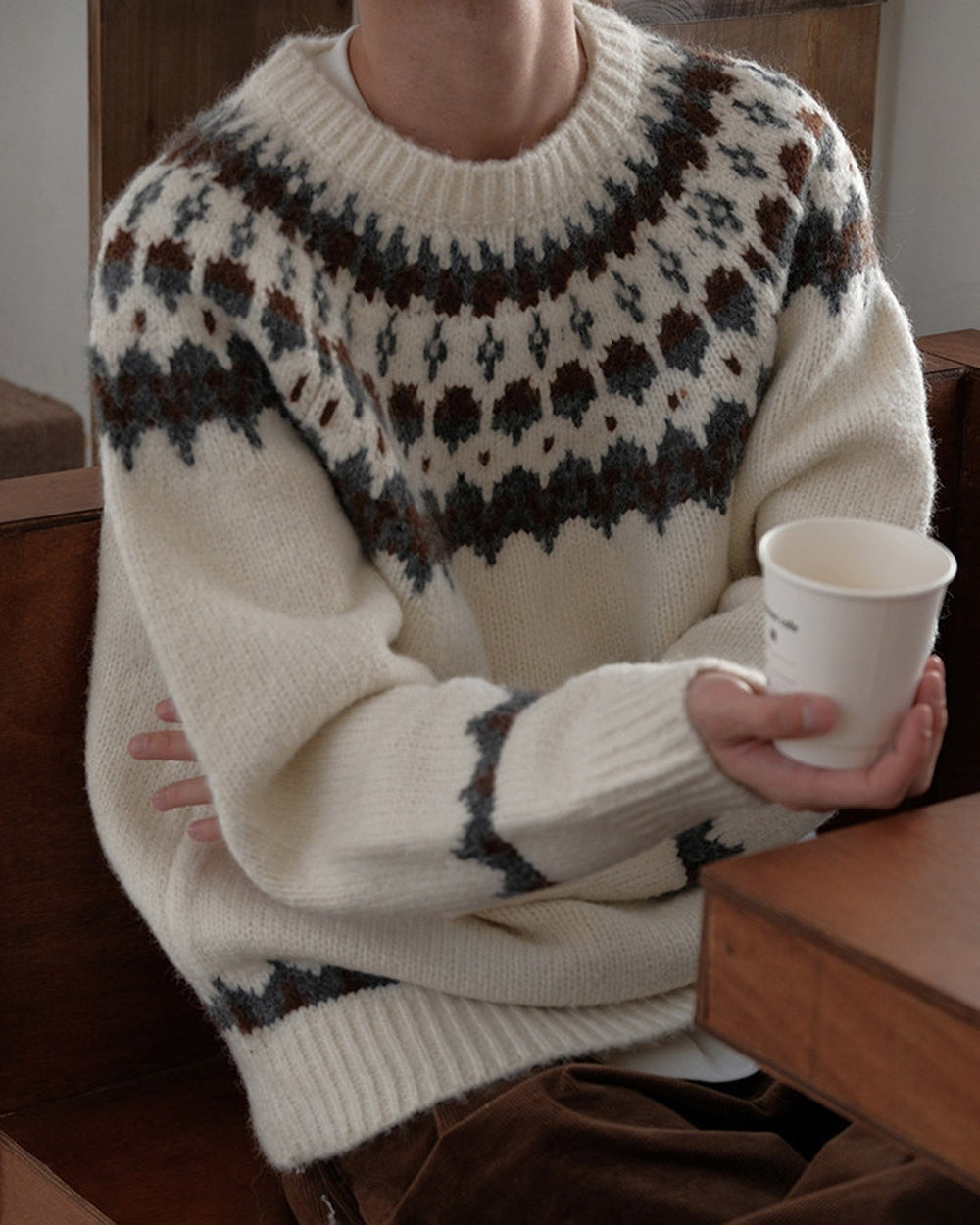 Man seated at a table in the cream fair-isle sweater, holding a paper cup