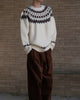 Man standing in a cream fair-isle yoke sweater with brown corduroy trousers, torso to knee