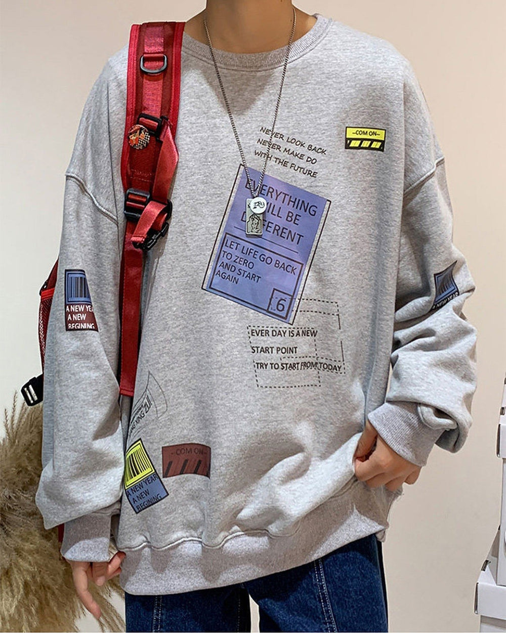 Light gray crewneck sweatshirt worn with a red backpack strap, showing the blue label print