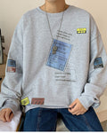 Man seated in the light gray crewneck sweatshirt, showing the blue label and the tags near the hem