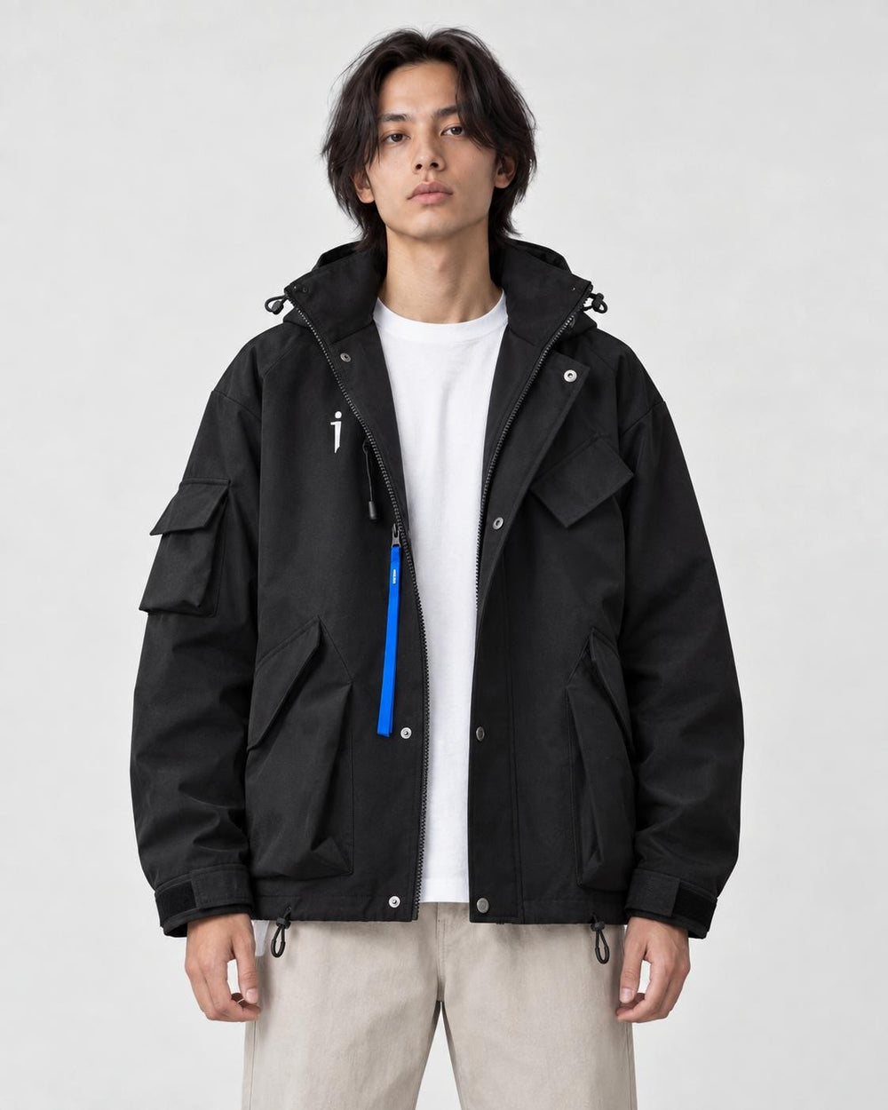 Man in a black hooded techwear jacket with a blue zip pull over a white tee and stone trousers, torso to mid-thigh
