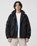 Man in a black hooded techwear jacket with a blue zip pull over a white tee and stone trousers, torso to mid-thigh