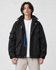 Man in a black hooded techwear jacket with a blue zip pull over a white tee and stone trousers, torso to mid-thigh