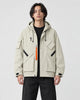Man in a beige hooded techwear jacket with an orange zip pull over a black tee, torso to mid-thigh
