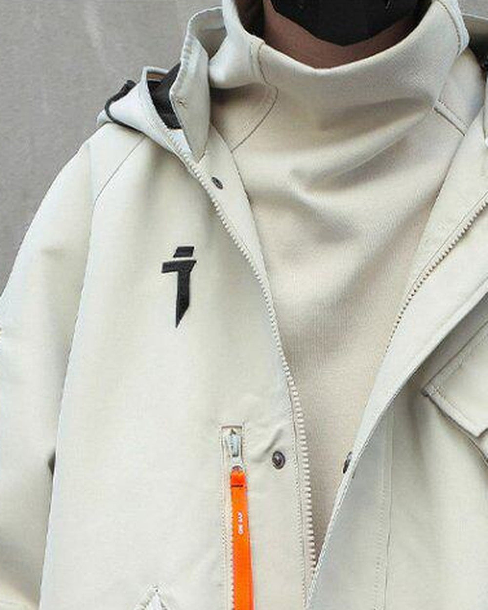 Close view of the beige jacket with the high collar, the black logo and the orange zip pull