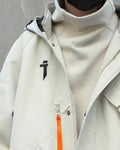 Close view of the beige jacket with the high collar, the black logo and the orange zip pull