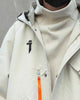 Close view of the beige jacket with the high collar, the black logo and the orange zip pull