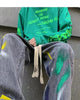 Man seated in the washed gray paint-splatter jeans, showing the green and yellow paint on the thigh