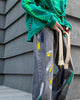 Washed gray paint-splatter jeans with two long frayed cotton tapes, worn with a green sweatshirt
