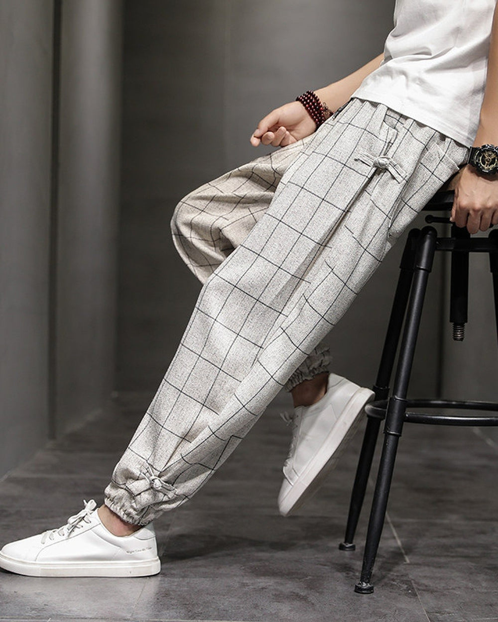 Man seated on a stool in the cream plaid harem pants, showing the frog-button knots at the hip and cuff