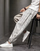 Man seated on a stool in the cream plaid harem pants, showing the frog-button knots at the hip and cuff