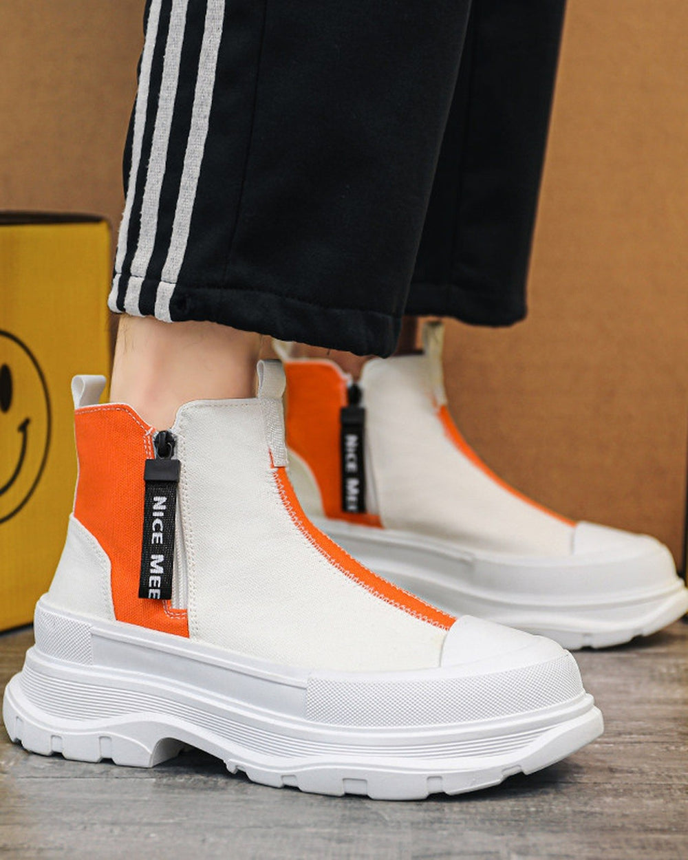 Orange and white platform sneaker boots on foot with black striped track pants, three-quarter view