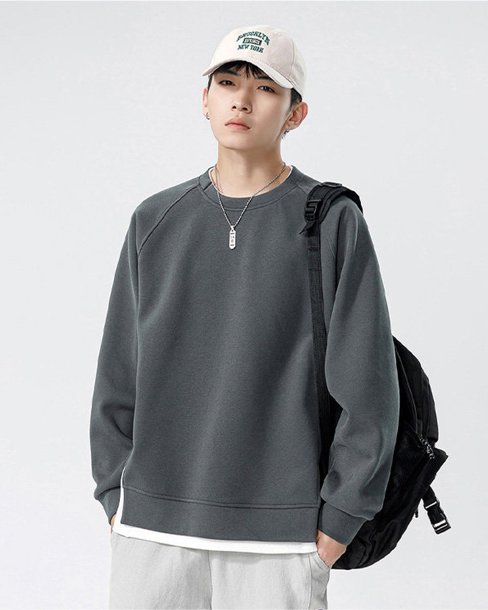 Dark gray raglan sweatshirt worn with a black backpack, showing the side slit and the white layered hem