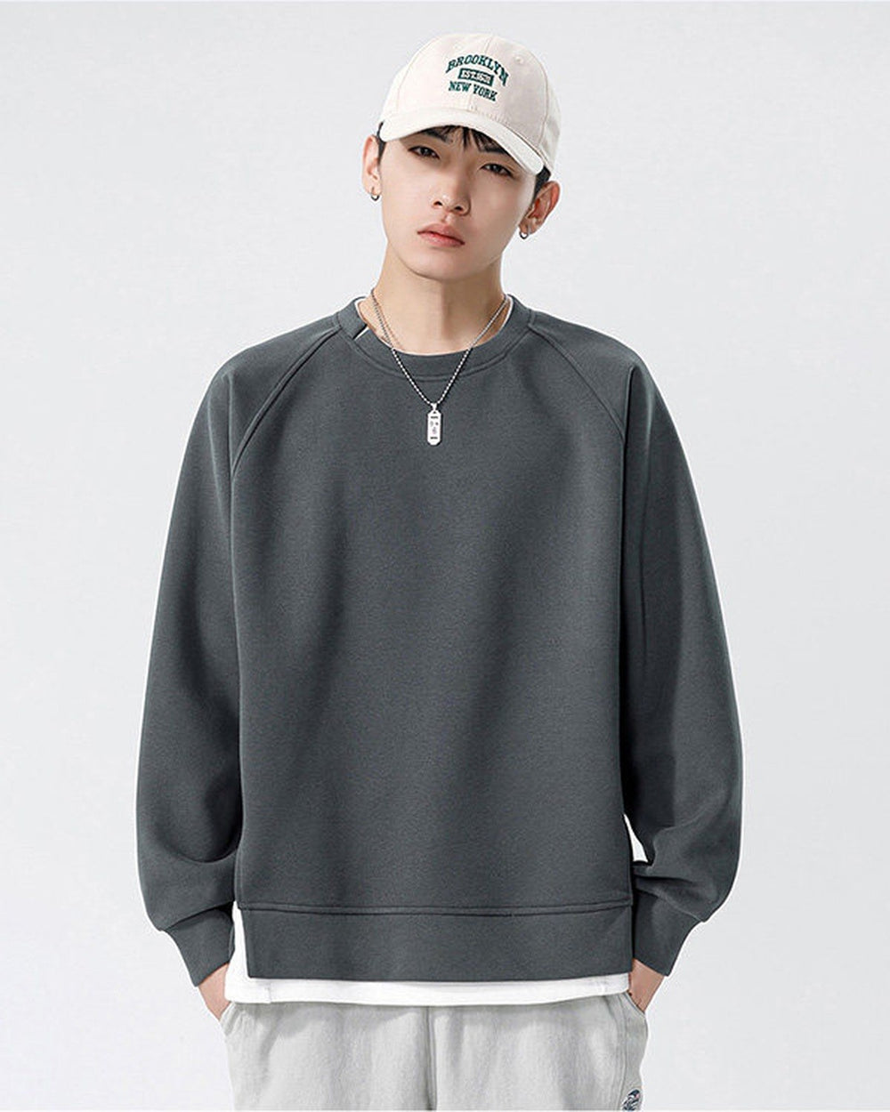 Dark gray raglan sweatshirt with a white layer showing below the hem, worn with a white cap and gray pants