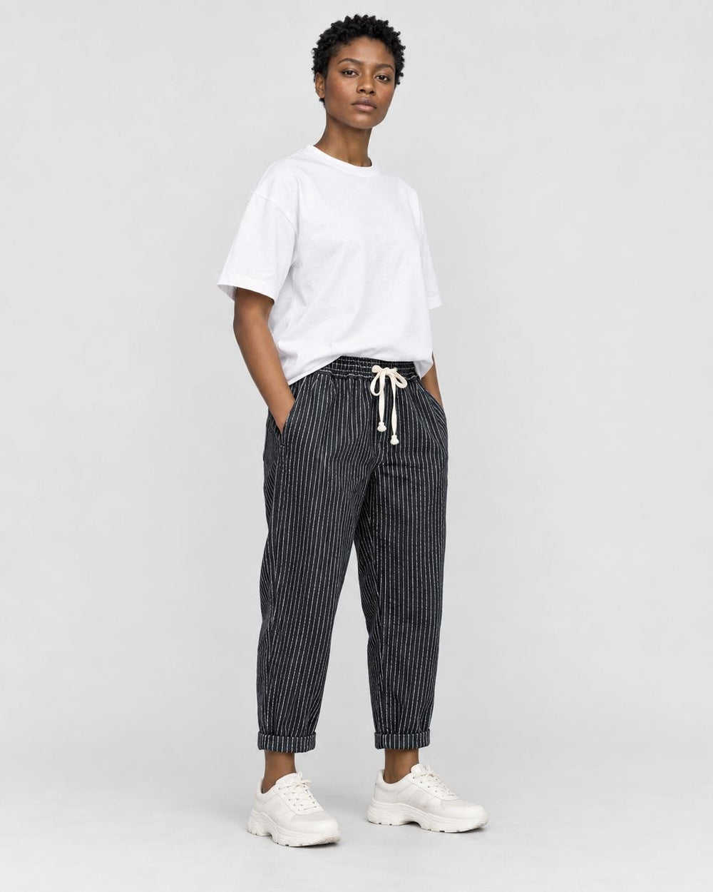 Model wearing the black striped cropped pants with a white tee and off-white sneakers, three-quarter view