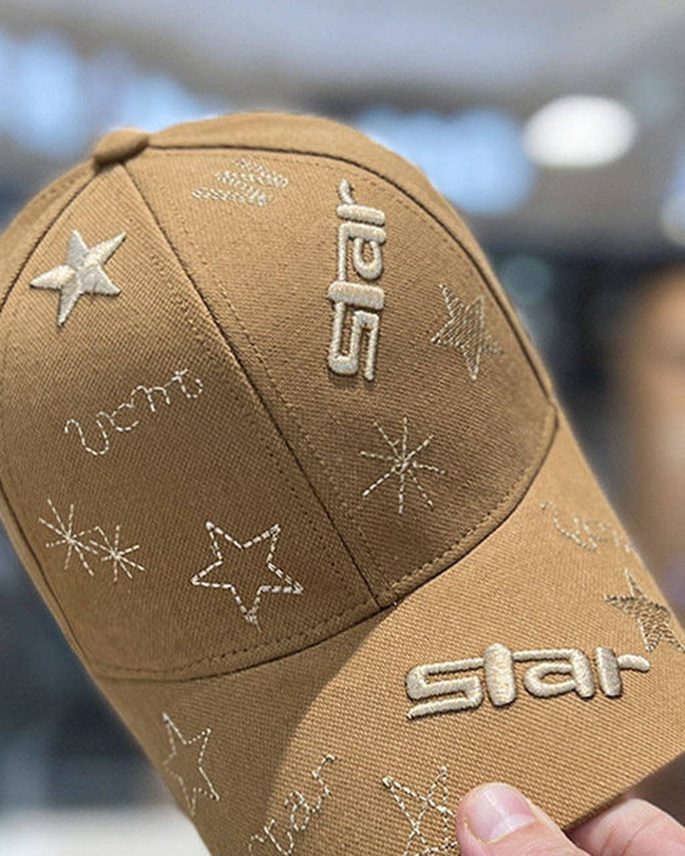 Tan-brown baseball cap held in a hand, with champagne star embroidery and the word star on the brim