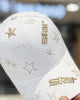 White baseball cap held in a hand, with gold star embroidery and the word star on the brim