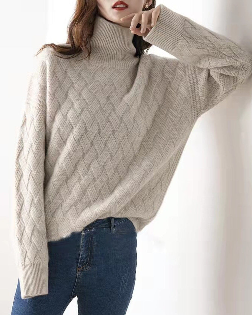 Woman leaning on a white wall in the beige woven sweater, one arm raised, the knit texture visible on the sleeve