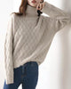 Woman leaning on a white wall in the beige woven sweater, one arm raised, the knit texture visible on the sleeve