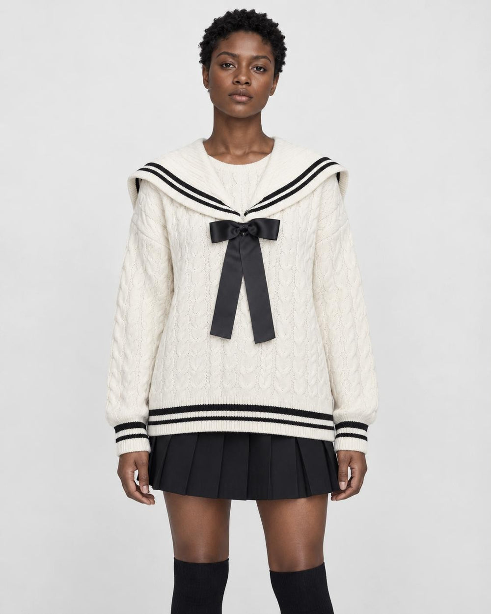Woman in a cream cable-knit sailor sweater with a black bow and striped collar over a black pleated skirt