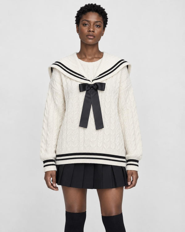 Woman in a cream cable-knit sailor sweater with a black bow and striped collar over a black pleated skirt