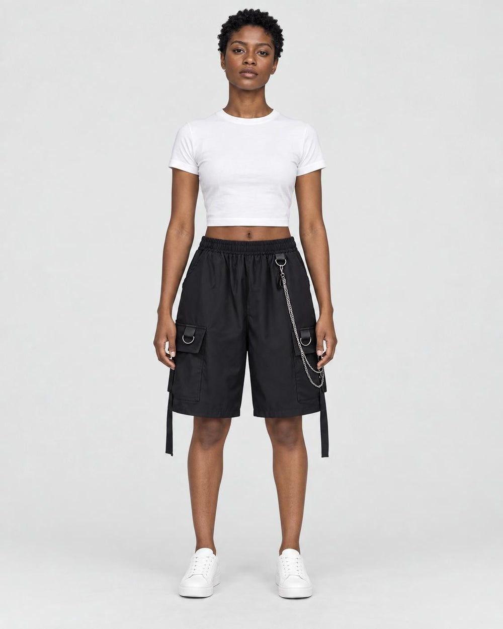Woman in black knee-length cargo shorts with a silver chain and side straps, a white cropped tee and white sneakers