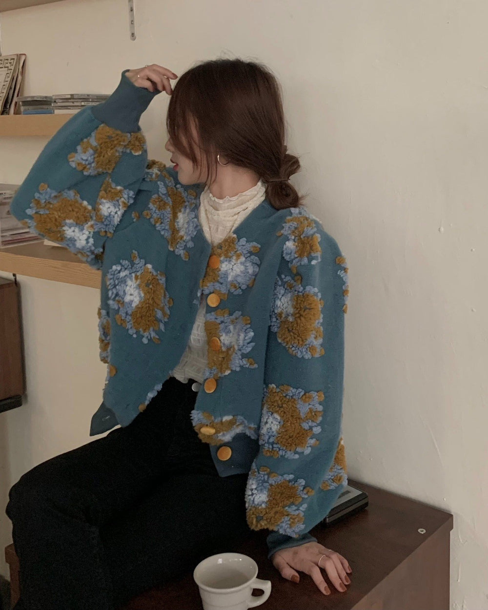 Woman seated in the teal flower-patch jacket with one hand raised to her hair