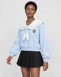 Woman in a light blue sailor-collar sweatshirt with an ivory collar and tie over a black pleated skirt, turned slightly