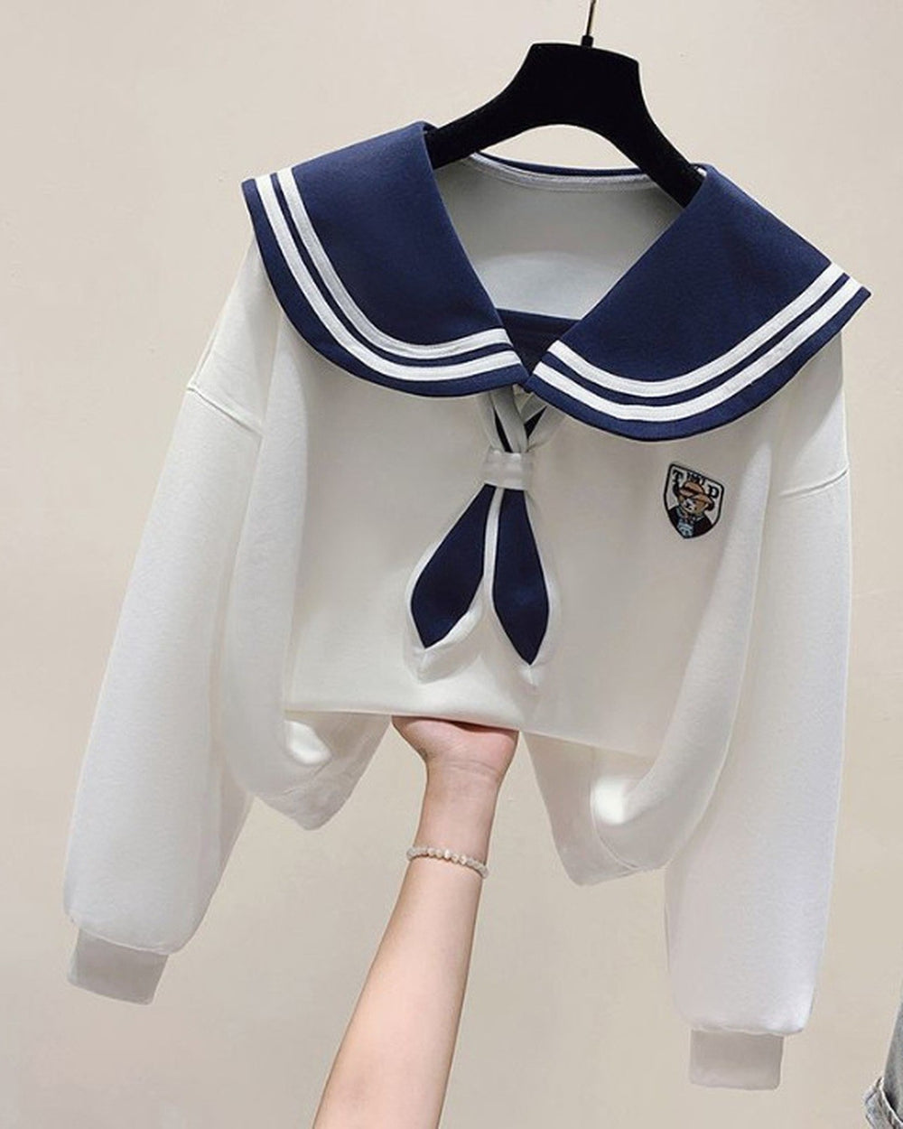 White cropped sweatshirt with a navy sailor collar striped in white, a navy tie and a crest, held up on a hanger