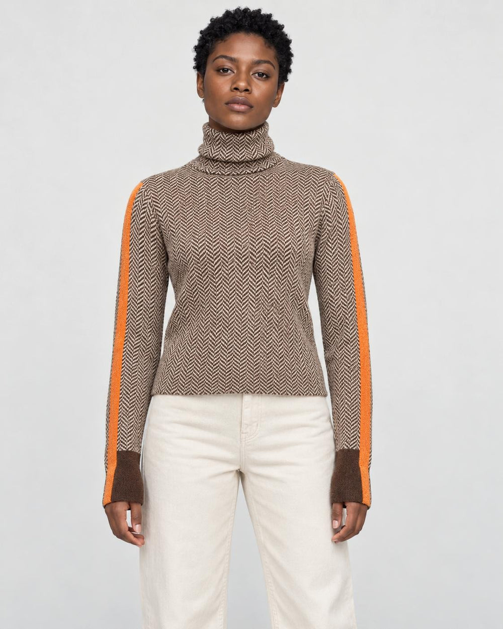 Woman standing in the herringbone turtleneck with orange sleeve stripes and cream trousers, front view