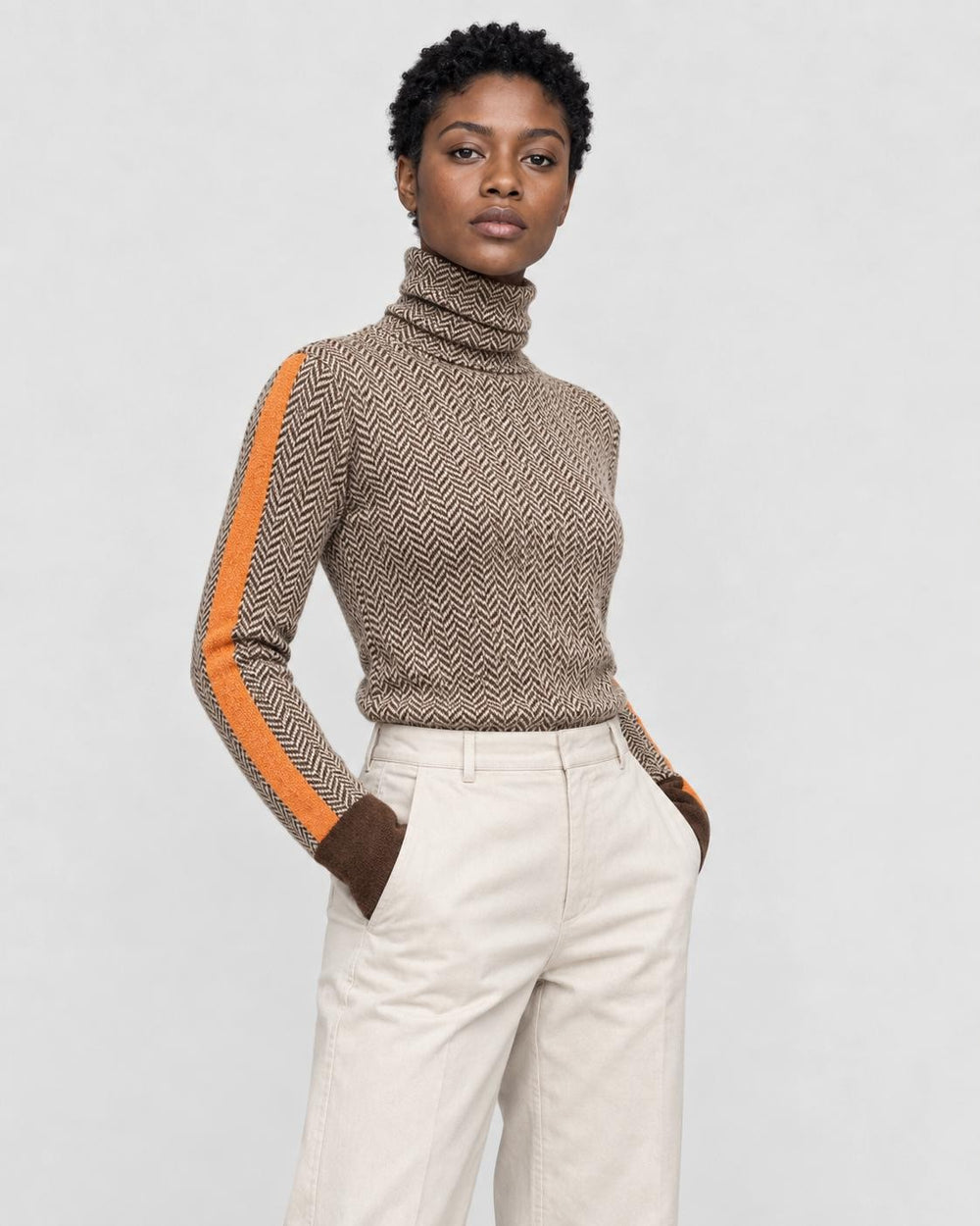 Woman in a brown herringbone turtleneck with an orange sleeve stripe and cream trousers, three-quarter view