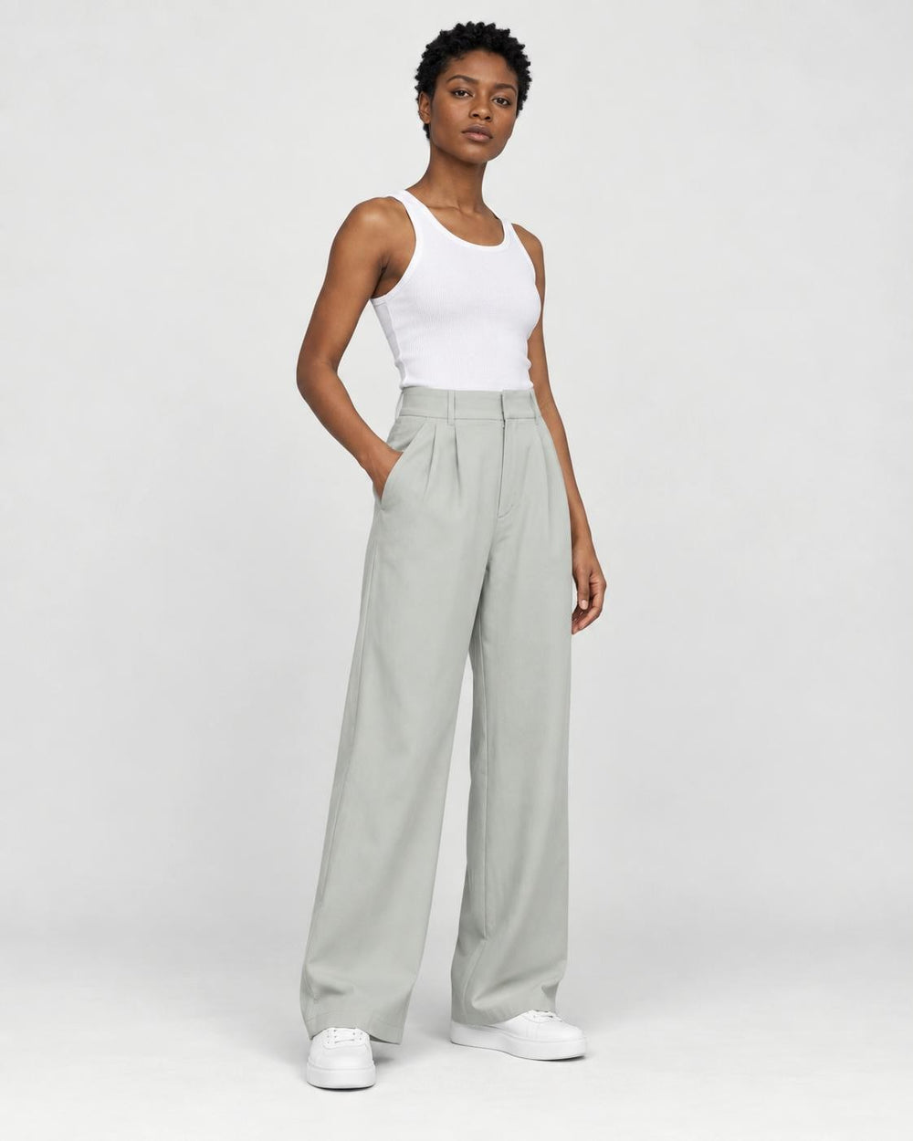 Model wearing the sage green high-rise pleated wide-leg pants with a white tank top and white sneakers, three-quarter view
