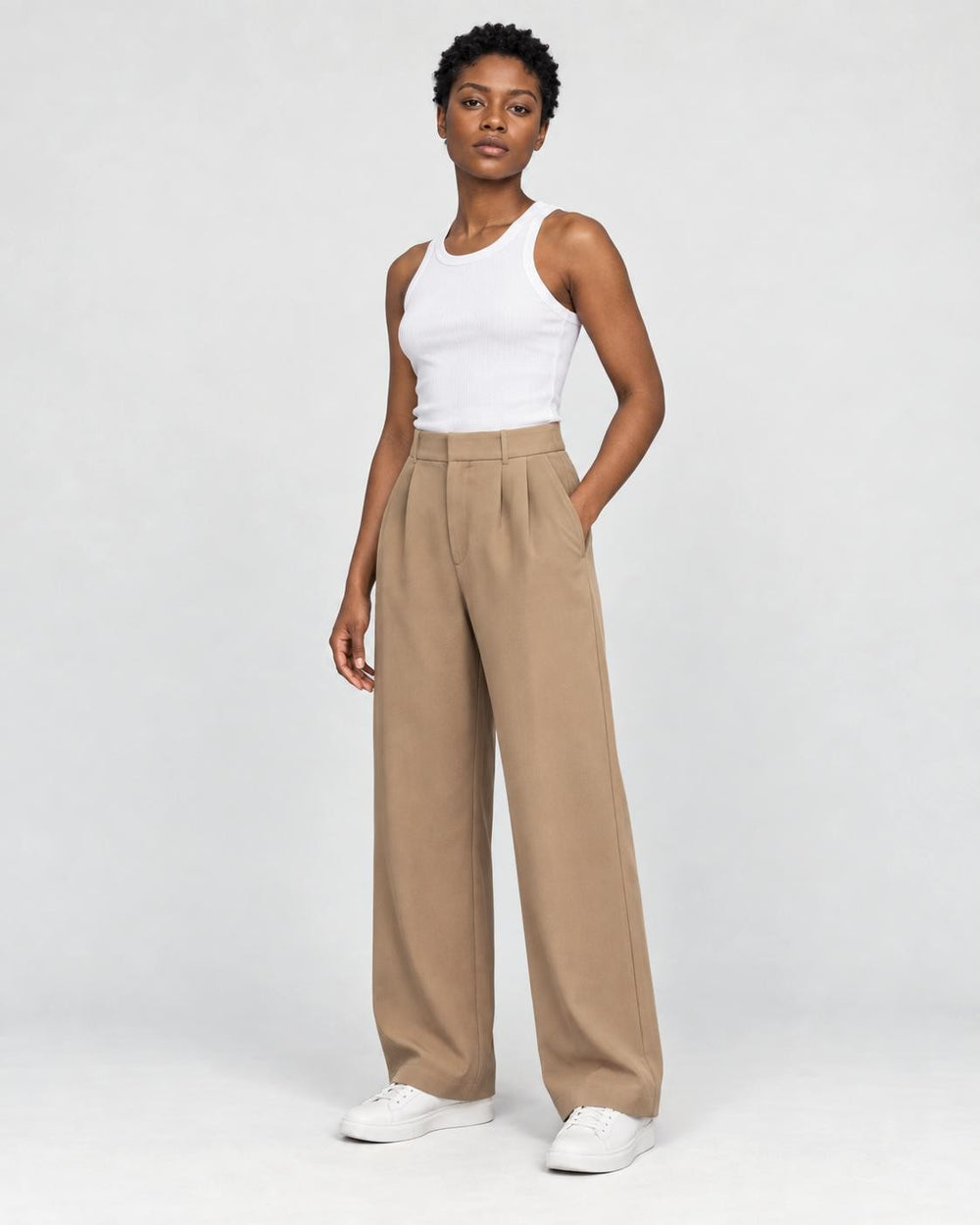 Model wearing the tan high-rise pleated wide-leg pants with a white tank top and white sneakers, three-quarter view